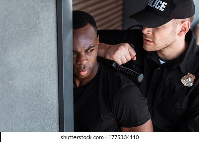 selective focus of police officer in uniform and cap holding truncheon while arresting african american man