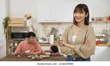 Selective Focus Portrait Of Confident Asian Mom Looking At Camera With Smile And Folded Arms As The Dad And Son Talking And Eating At Dining Table At Background