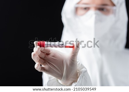 selective focus of positive covid-19 omicron variant test in hand of blurred microbiologist in hazmat suit isolated on black