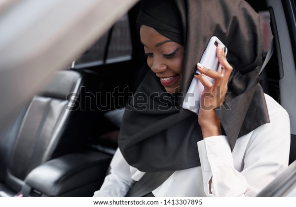 Selective\
focus of pretty islamic girl wearing black hijab sitting in big car\
and talking by phone. Muslim woman looking down and laughing while\
driving. Concept of automobile and\
comfort.