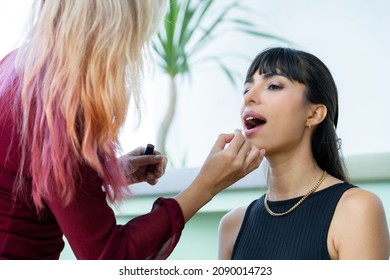 Selective Focus Of A Professional Female Make-up Artist's Hand Carefully Applied Liquid Lipstick On Lips Of A Young Beautiful Caucasian Model With Bangs Sitting Getting Make-up In A Salon Or Studio.