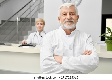 Selective Focus Of Professional Therapist Posing With Arms Crossed And Looking At Camera. Portrait Of Eldery Caucasian Male Doctor In White Lab Coat, Woman Behind Reception On Background.