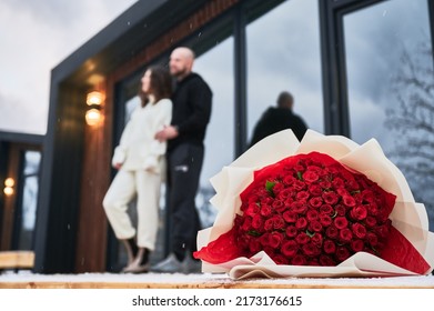 Selective Focus Of Red Roses Bouquet With Couple In Love On Blurred Background. Man And Woman Standing Outside Scandinavian House Barnhouse With Flowers On Forefront.