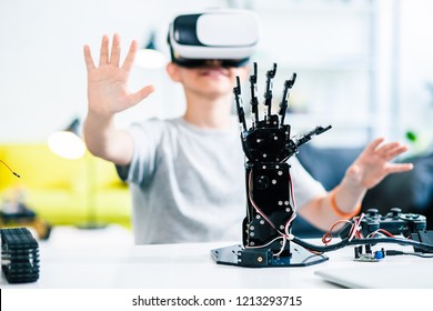 Selective focus of a robotic hand on the table with a little smiling boy testing VR glasses in the background
