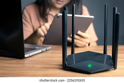 Selective focus at router. Internet router on working table with blurred happy woman using tablet at the background. Fast and high speed internet connection from fiber line with LAN cable connection. - Shutterstock ID 2256192655