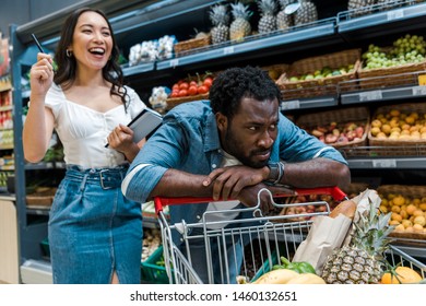 selective focus of sad african american man near shopping cart near happy asian woman with notebook
