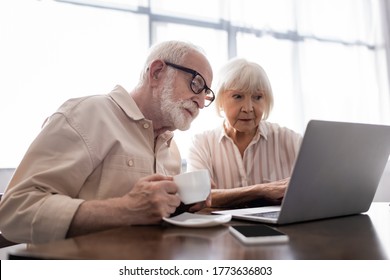 Selective focus of senior man drinking coffee while wife using laptop near smartphone on table - Powered by Shutterstock