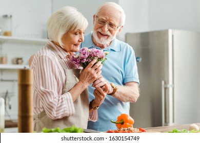 Selective focus of senior woman smelling bouquet beside smiling husband and vegetables on kitchen table - Powered by Shutterstock