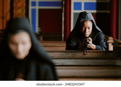 Selective focus shot of ethnically diverse young and mature Catholic nuns sitting on wooden benches in church and praying