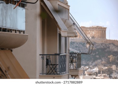 A selective focus shot looking through a modern apartment balcony towards the blurred background of the ancient Acropolis hill. The foreground features typical architectural details like awnings and wrought-iron railings, creating a sense of living in Athens with a view of history. Ideal for travel blogs, hotel promotion, or real estate listings
