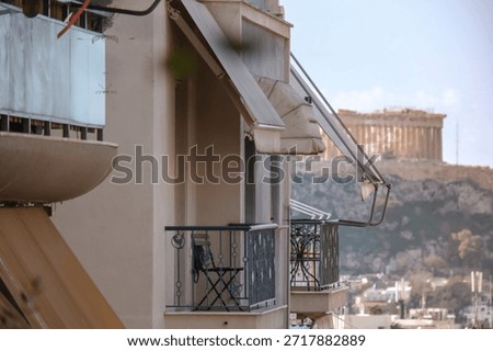 A selective focus shot looking through a modern apartment balcony towards the blurred background of the ancient Acropolis hill. The foreground features typical architectural details like awnings and wrought-iron railings, creating a sense of living in Athens with a view of history. Ideal for travel blogs, hotel promotion, or real estate listings
