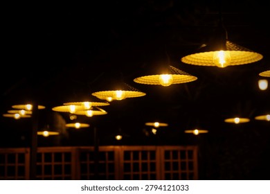 A selective focus shot of multiple decorative straw-woven lampshades with burning light bulbs illuminating a dark outdoor terrace or restaurant setting at night.