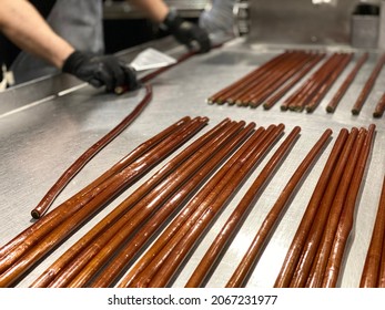 Selective Focus Shot On Hard Candy Making Process. Rolls Of Brown Kiwi Shaped Hard Candy On Stainless Steel Table. Man With Black Gloves Getting Ready To Cut Candy With Metal Spatula In Background.