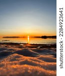 Selective focus shot of a sunset in Helsinki, in front of the gulf of Filand. Snowy beach in Finland with orange sky. Shot of a beach in baltic sea covered with snow. Shiny snow with reflection.