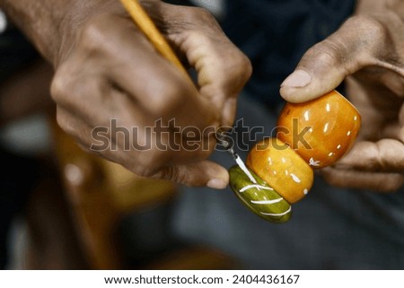 A selective focus of a small and simple colorful wooden toy being painted with a brush at a workshop in Channapatna, Karnataka, India. The Indian man's hand is out of focus.