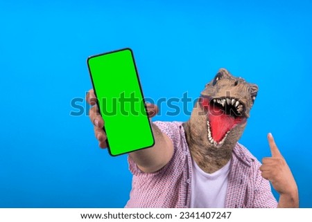 selective focus of smartphone with green screen being held by man with T-Rex head on blue background
