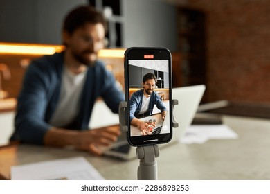 Selective focus of smartphone on tripod recording live video of successful businessman vlogger coach teching online to camera streaming for podcast giving business class presentation training