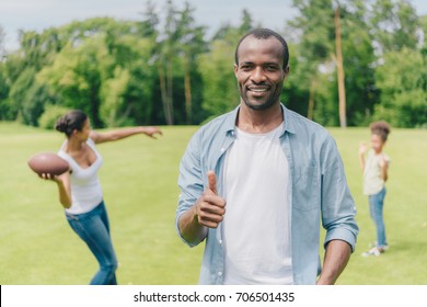 Selective Focus Of Smiling African American Man Showing Thumb Up While Family Playing American Football Behind