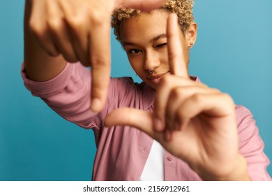 Selective focus of smiling black girl with blonde curly hair and piercing making frame with hands and fingers, looking through it, closing one eye. Creativity and photography concept. Body language - Shutterstock ID 2156912961