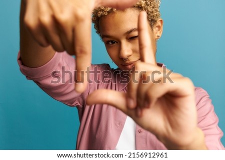 Selective focus of smiling black girl with blonde curly hair and piercing making frame with hands and fingers, looking through it, closing one eye. Creativity and photography concept. Body language