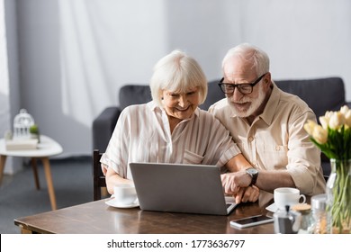 Selective focus of smiling elderly couple using laptop near coffee cups on table - Powered by Shutterstock