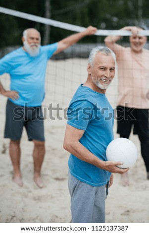selective focus of smiling elderly man with volleyball ball looking at camera with old friends behind on beach