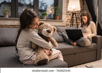 selective focus of smiling kid hugging teddy bear and mother working on laptop at home