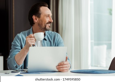 Selective focus of smiling man drinking coffee and using laptop on kitchen table - Powered by Shutterstock