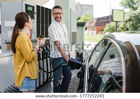 Selective focus of smiling man looking at wife with coffee to go while refueling car on gas station