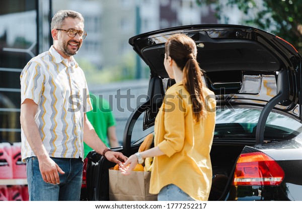 Selective focus of smiling man looking at woman\
near open car trunk on urban\
street