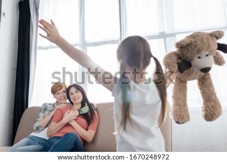 Selective focus of smiling mothers looking at daughter with teddy bear in living room