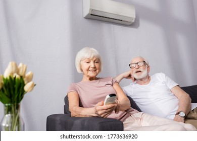 Selective focus of smiling senior woman holding remote controller of air conditioner near husband at home
