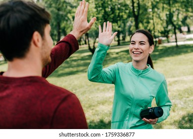 Selective focus of smiling sportswoman with sports bottle high five to boyfriend in park - Shutterstock ID 1759336877