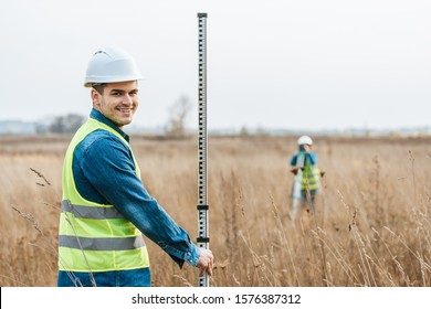 Selective focus of smiling surveyor with ruler and colleague with digital level in field