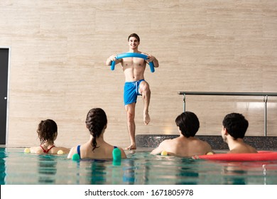 Selective focus of smiling trainer with pool noodle training with group of people in swimming pool