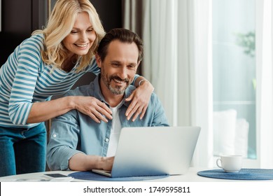 Selective focus of smiling woman embracing husband using laptop in kitchen