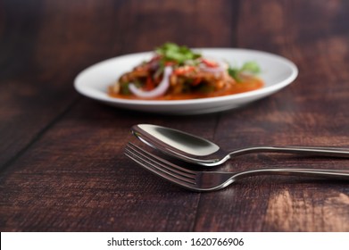 Selective Focus Stainless Spoon And Fork On Wooden Table, Blured Background With Spicy Salad Of Sardine With Tomato Sauce In White Dish, Copy Space