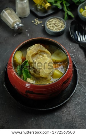 Selective focus of Sup Kikil (beef tendon soup) - Shiny, Oily and Fatty of Indonesian beef tendon soup with vegetables served in red bowl over dark textured backdrop with ingredients