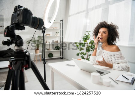 selective focus of surprised african american influencer in braces applying cosmetic cream near digital camera