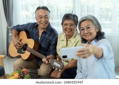 Selective focus, Three happy senior friends gathered indoors, one playing acoustic guitar while others sing animatedly, female friend use smartphone selfies with camaraderie in living room - Powered by Shutterstock