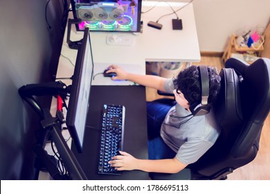 Selective focus to top view of Asian boy gamer playing games on computer in the room at home, wearing headphones and using backlit colorful keyboard on wooden table.