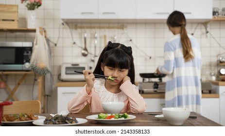 selective focus of unhappy picky Asian schoolgirl daughter holding face with disgust look on face while playing with food in bowl at dining table. her mother is cooking in kitchen at background - Powered by Shutterstock