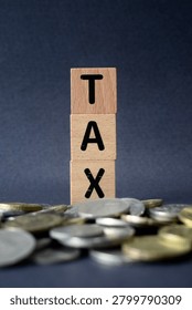 A selective focus vertical macro shot of wooden blocks spelling TAX emerging from a blurred heap of silver coins, signifying tax burdens, financial savings, and microeconomics.