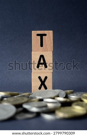 A selective focus vertical macro shot of wooden blocks spelling TAX emerging from a blurred heap of silver coins, signifying tax burdens, financial savings, and microeconomics.