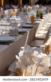 Selective focus vertical shot of white delicate flowers on a chair in the foreground with a long decorated outdoor wedding banquet table blurred in the background. Elegant event decoration, romantic celebration and catering concept.