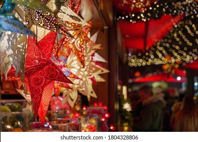 Selective Focus View Of Star Cut-out Paper Hanging Lantern In Front Of Stall During Christmas Market In Germany With Blur Background With Bokeh Of Illuminated Night Atmosphere.