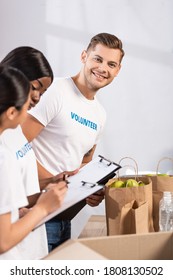 Selective focus of volunteer looking at camera near multiethnic women with clipboards and donations