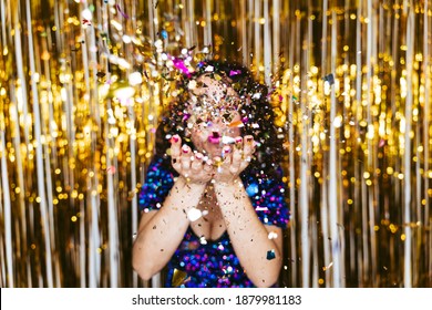 Selective focus. A woman in an elegant shiny dress and Christmas decorations behind her blowing some confetti to the camera. New Year's Eve party concept - Powered by Shutterstock