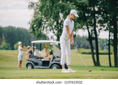 selective focus of woman with golf club playing golf and friends resting at golf cart on green lawn