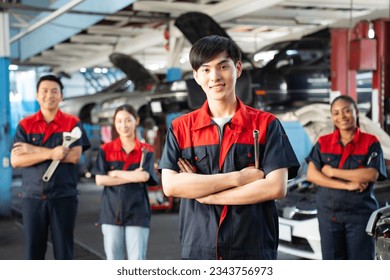 Selective focus of a young Asian male mechanic in uniform, standing to hold a wheel spanner with arms folded and smiling at camera with a blurred mechanic team standing in the background in the garage - Powered by Shutterstock